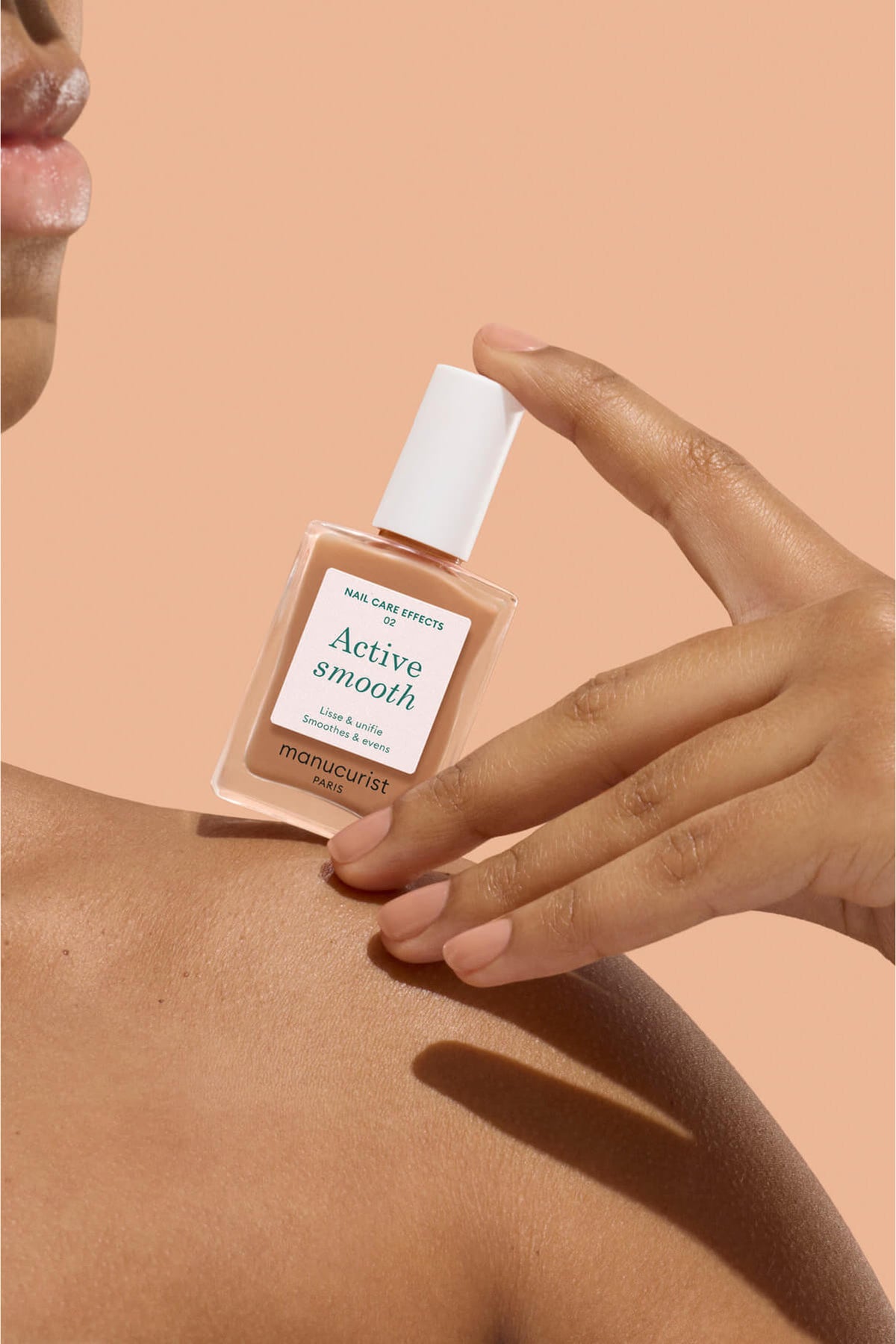 Une personne à la peau lisse tient un flacon de soin pour les ongles Manucurist étiqueté « Active Smooth » sur un fond couleur pêche. Ses ongles sont impeccablement manucurés et au rendu naturel.