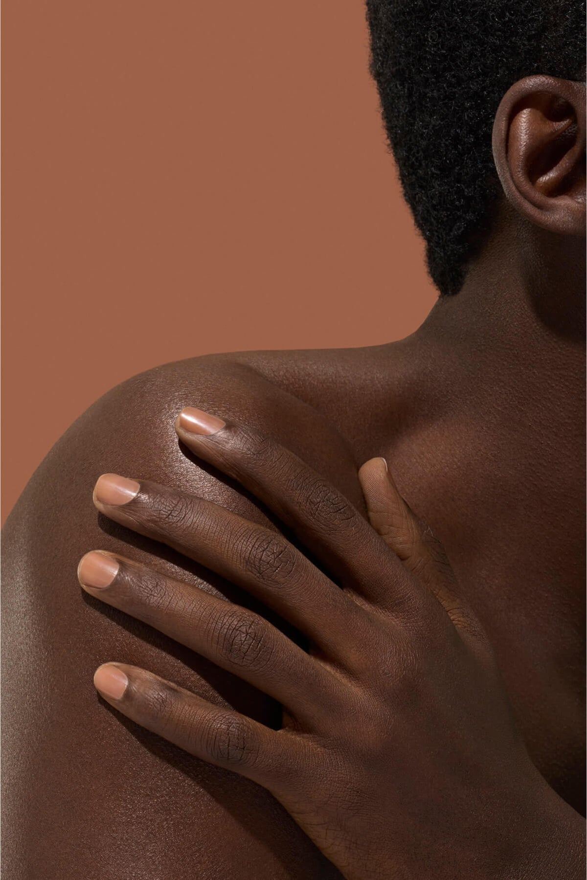 Gros plan d'une personne à la peau foncée touchant son épaule nue de la main, sur un fond marron doux. Seule une partie du visage, de l'épaule et de la main est visible.