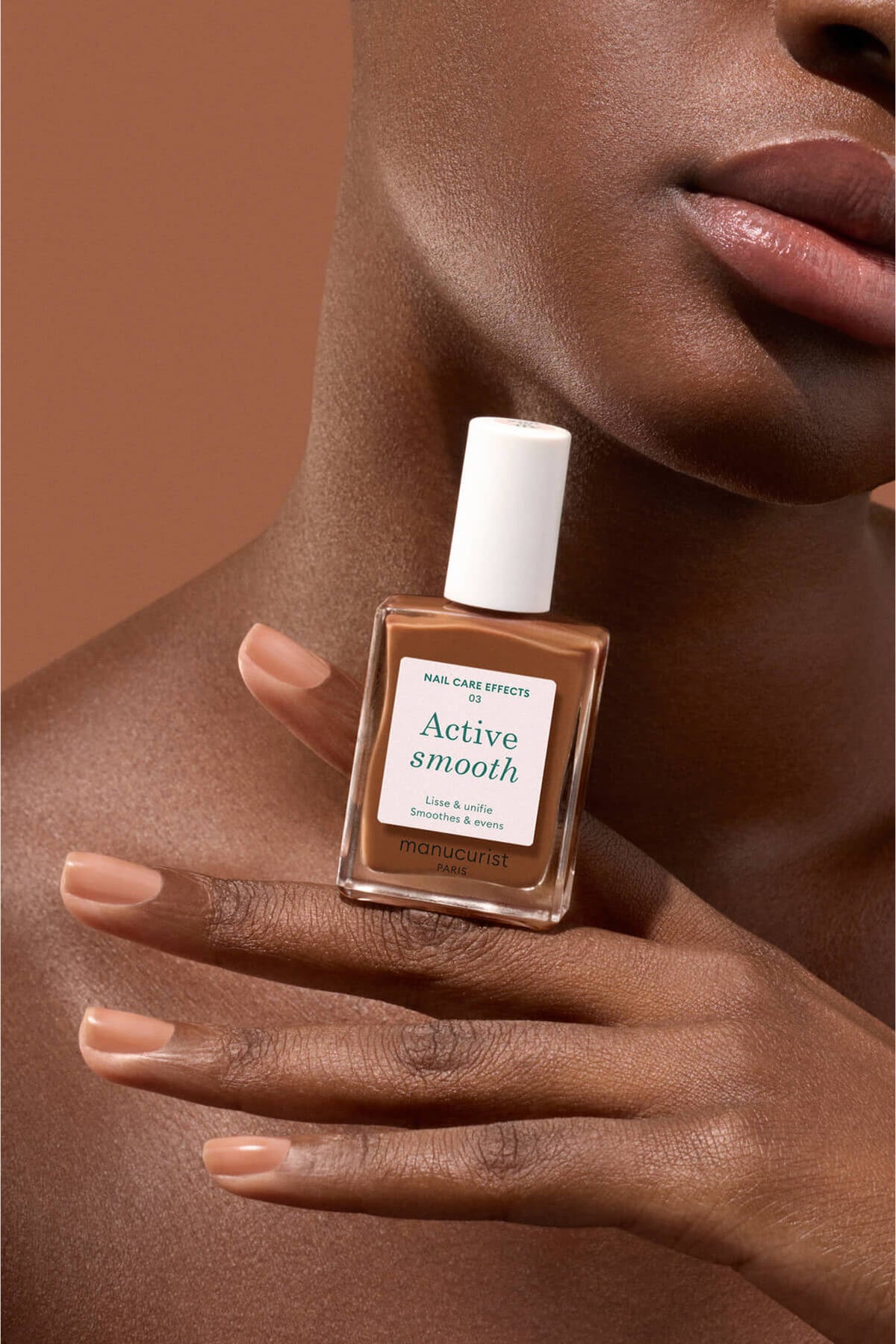 Une personne à la peau moyennement foncée tient un flacon de soin des ongles Manucurist Active Smooth, assorti à la couleur naturelle de ses ongles et de ses lèvres, sur un fond brun chaud.