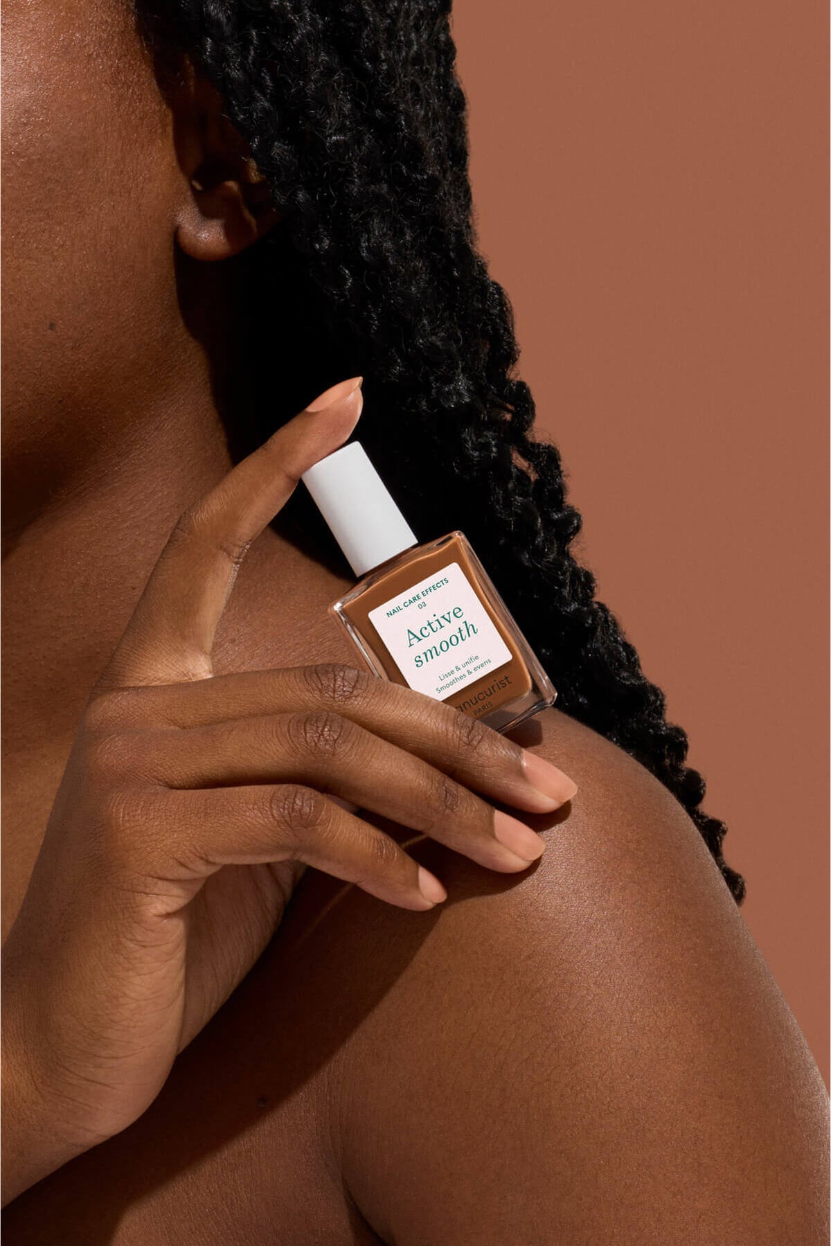 Une personne à la peau foncée et aux cheveux tressés tient un flacon de vernis à ongles intitulé Achieve Smooth près de son visage, sur un fond marron uni.