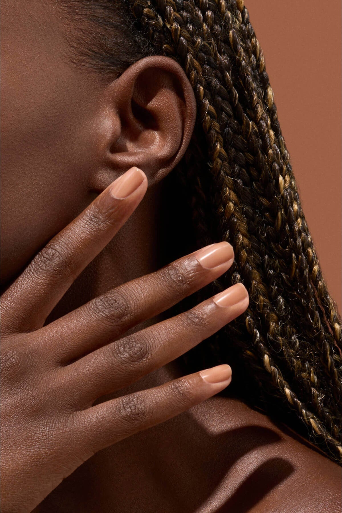 Gros plan d'une personne à la peau brun moyen et aux cheveux tressés touchant son visage, montrant des ongles soigneusement manucurés vernis dans une teinte nude sur fond marron.