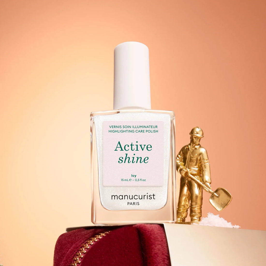 Un flacon de vernis à ongles Manucurist Active™ Shine Icy se trouve à côté d’une petite figurine dorée représentant un ouvrier du bâtiment tenant une pelle.