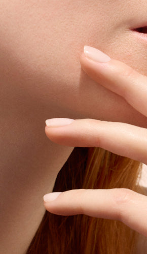 Gros plan sur le bas du visage et le menton d'une personne avec deux doigts touchant délicatement la peau. Les ongles sont impeccablement manucurés avec un vernis clair et naturel. L'image met en valeur une peau lisse et saine ainsi que des ongles soignés.
