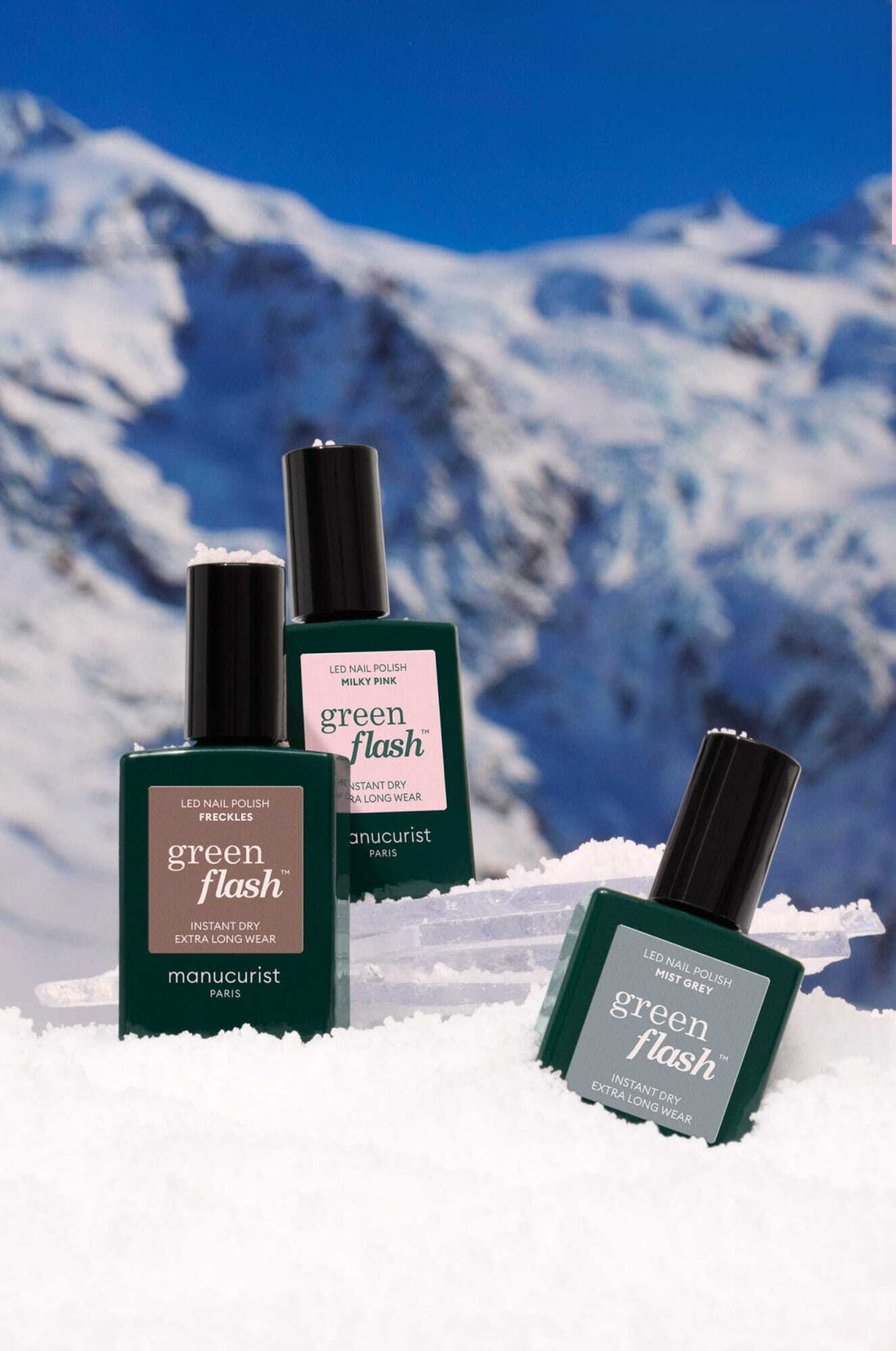 Three bottles of Manucurist Green Flash nail polish are displayed on snow with a backdrop of snowy mountains and a clear blue sky.