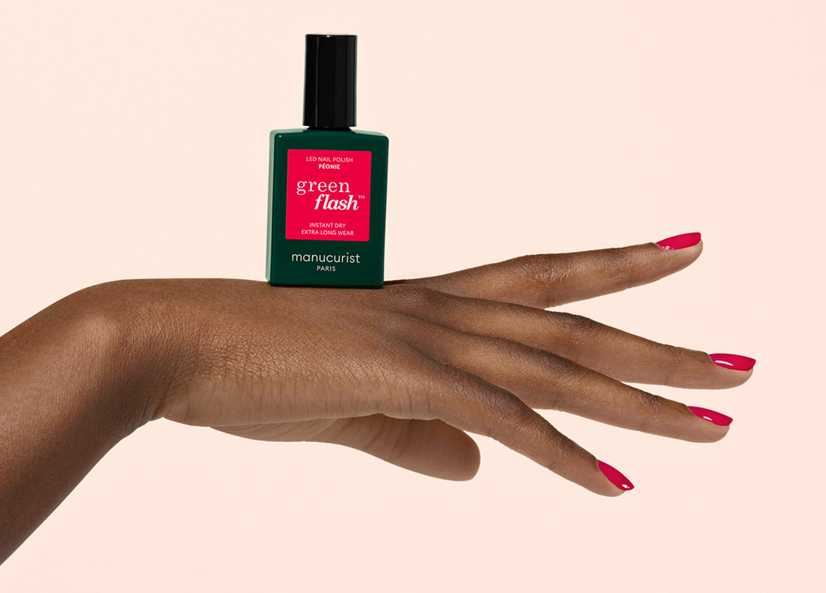 A hand with red-painted nails is held flat, balancing a green bottle of Manucurist Green Flash nail polish on top. The background is a plain, light beige color.