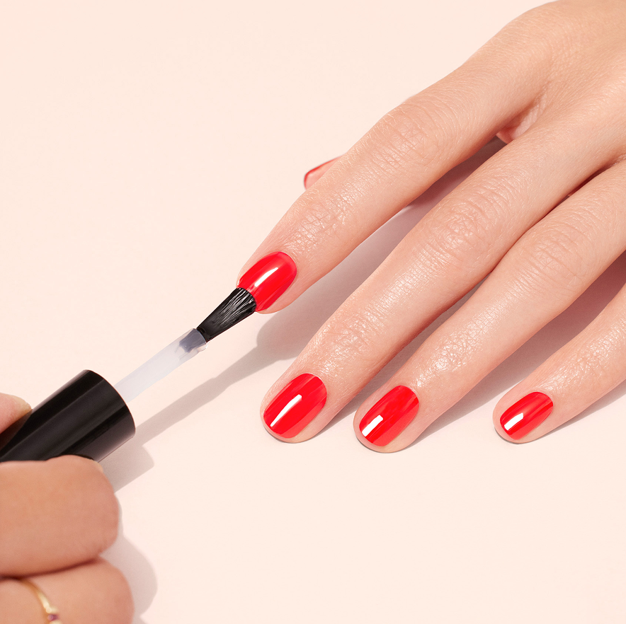 A hand is being painted with bright red nail polish by another hand using a brush. The nails are neatly manicured and the background is a soft, light peach color.
