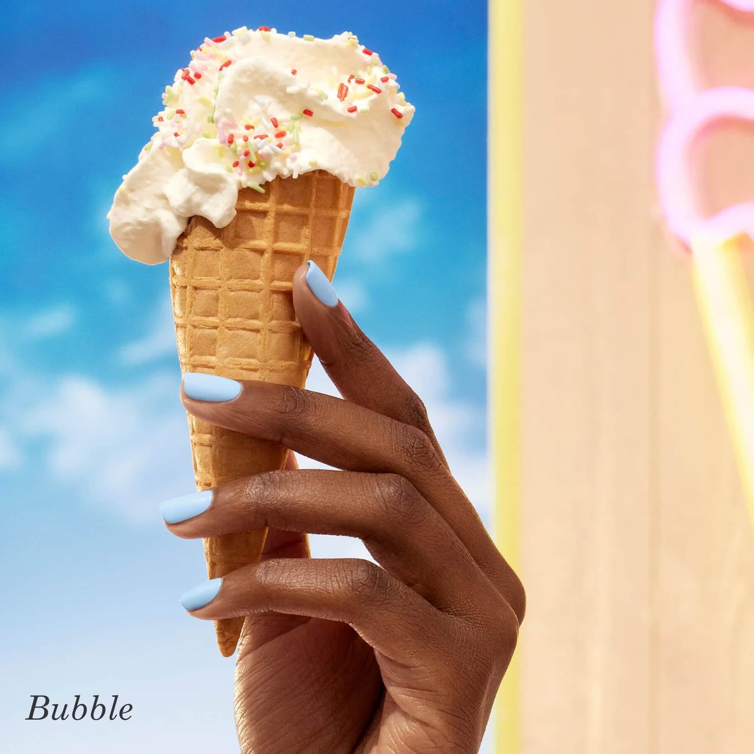 A hand with pastel blue nail polish holds a waffle cone topped with whipped cream and colorful sprinkles, against a bright blue sky background.