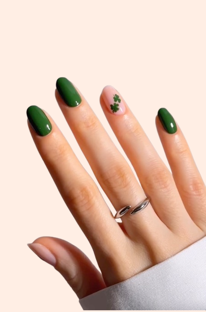 A hand with green polished nails; the ring finger has a nude base with two small green shamrock designs. The person wears a silver ring, and the background is light beige.