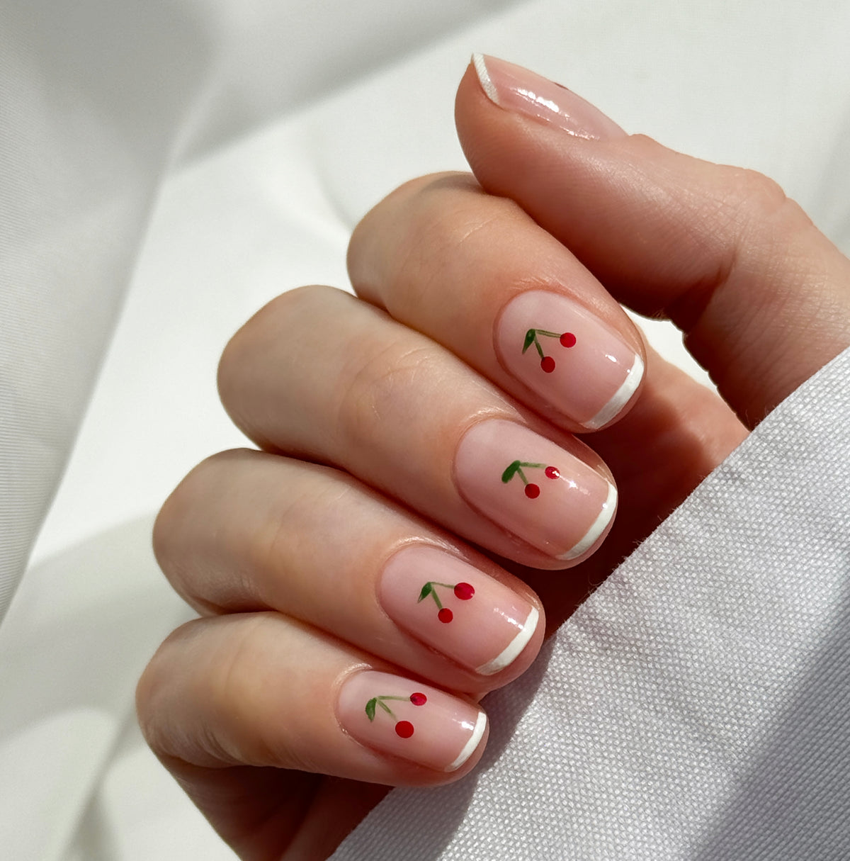 A hand with a French manicure featuring small red cherry designs with green stems painted on several nails, against a soft white background.