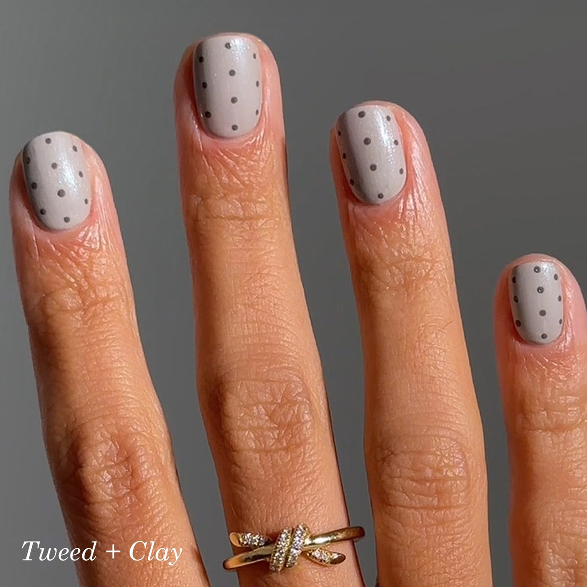 A hand with light brown nails featuring small black polka dots. The person wears a gold ring with a knot design on their ring finger. The background is neutral gray. Text in the corner reads Tweed + Clay.