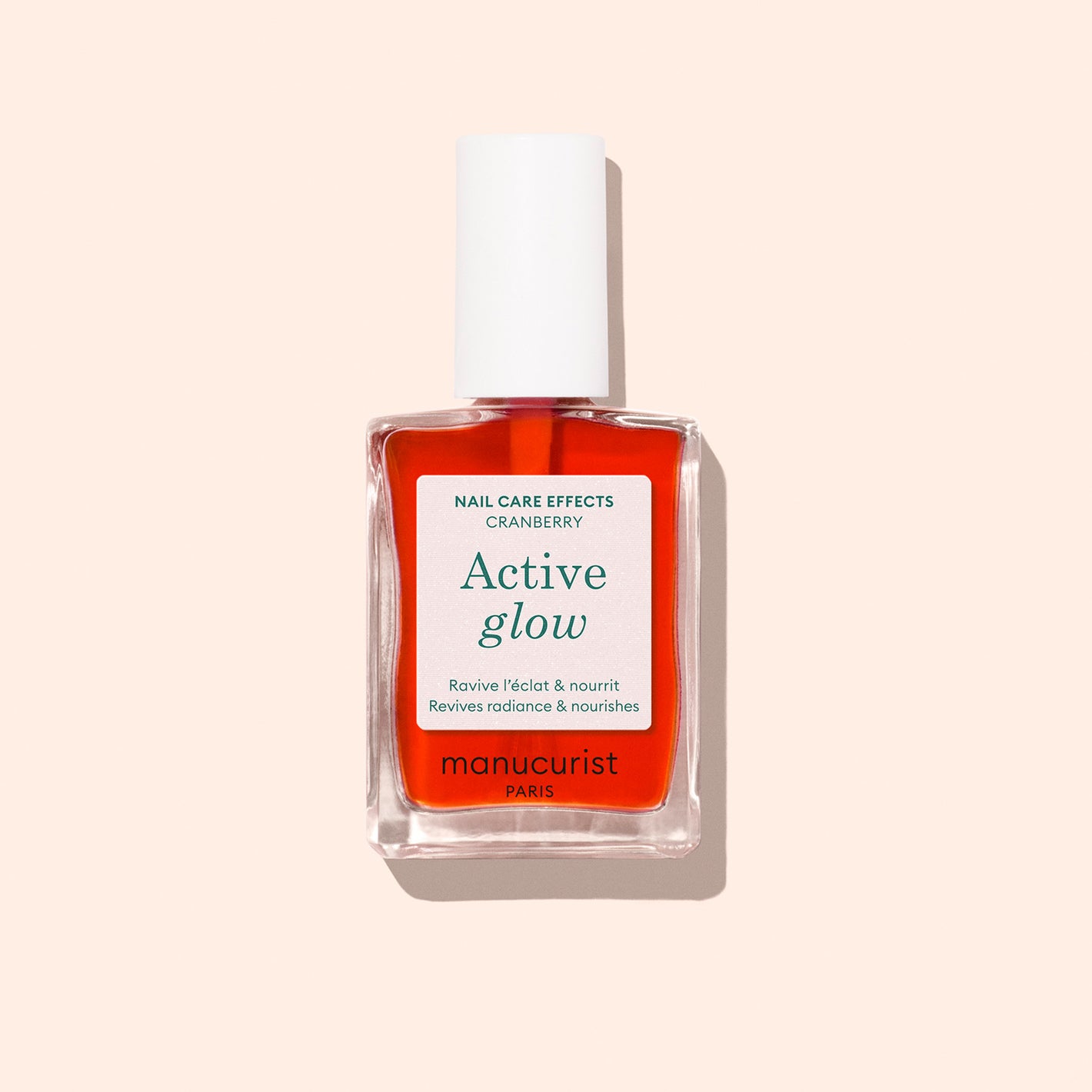 A bottle of Manucurist Active Glow Cranberry nail polish, featuring coral gloss-effect and a plant-based formula, with red liquid and a white cap, is shown against a light beige background.