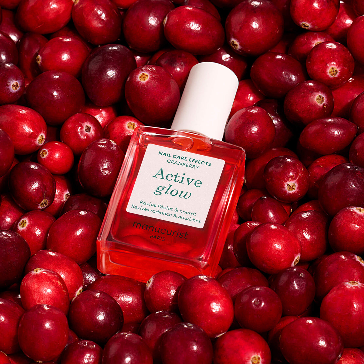 Un flacon carré d’huile de soin pour les ongles Manucurist Active™ Glow Cranberry, enrichie en ingrédients d’origine végétale, est présenté sur un lit de canneberges fraîches. L’étiquette met en avant la canneberge et ses bienfaits, comme raviver et nourrir les ongles, pour un éclat teinté et sain.