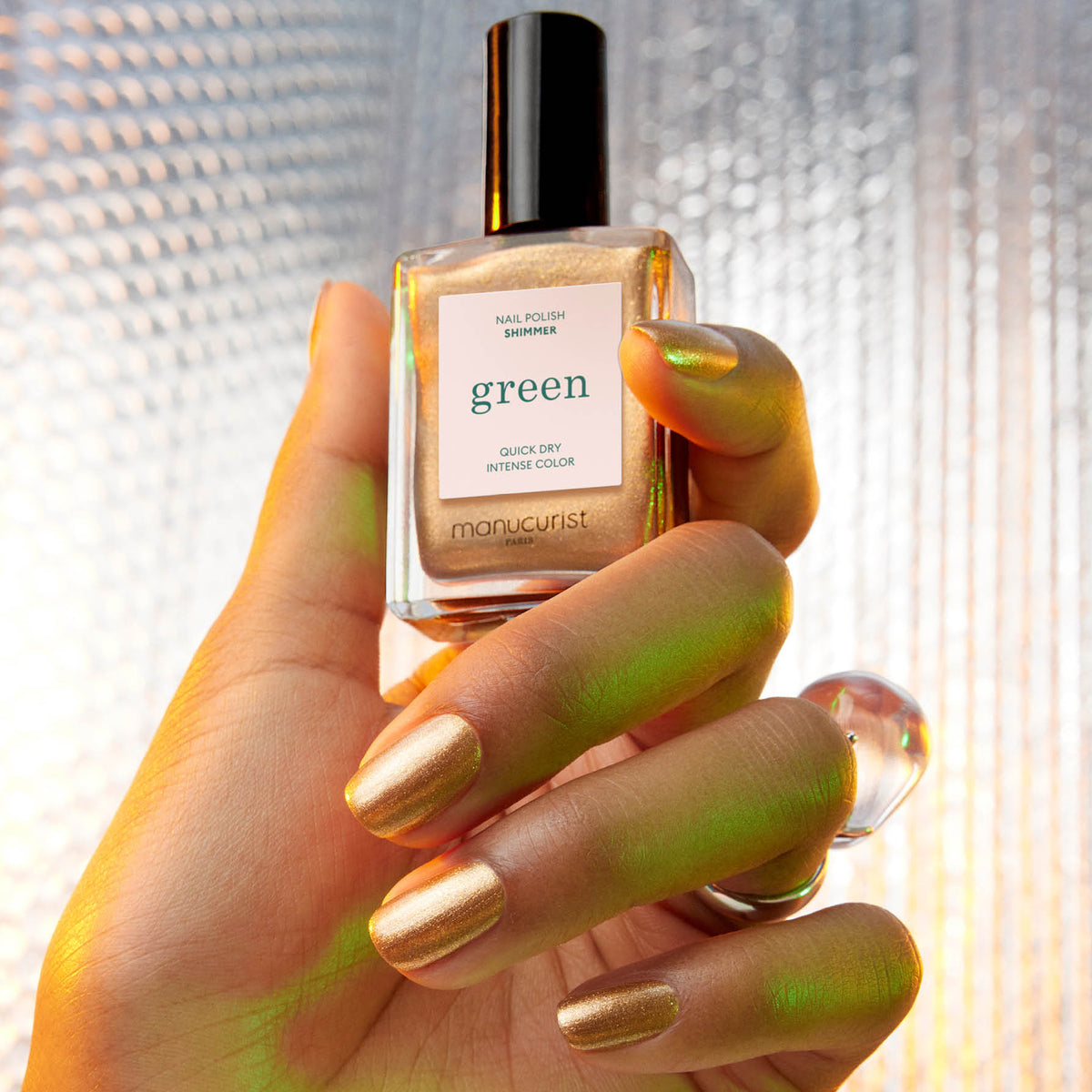 Une main portant un vernis à ongles doré métallisé scintillant de Manucurist tient un flacon assorti "Shimmer" de Manucurist sur un fond texturé et délicatement éclairé.
