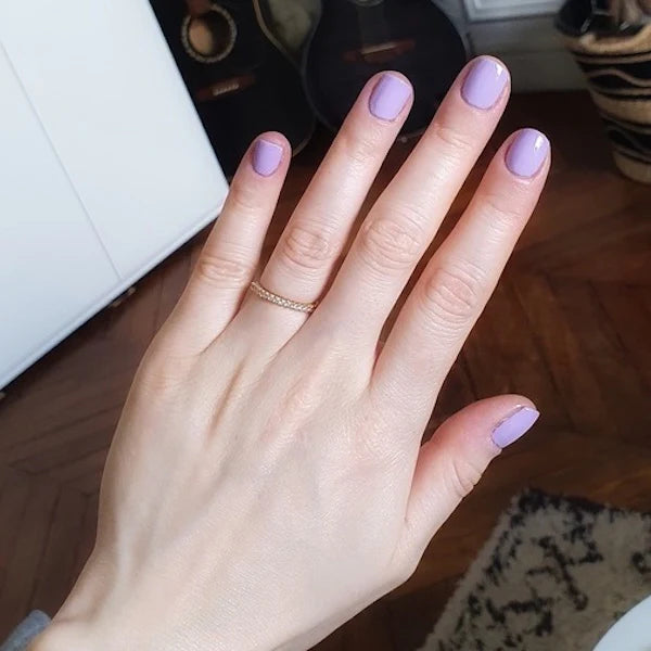 Une main à la peau claire aux ongles courts couleur lavande est présentée, portant une délicate bague en or. En arrière-plan, on aperçoit des guitares, un panier et une partie de tapis sur un sol en bois.