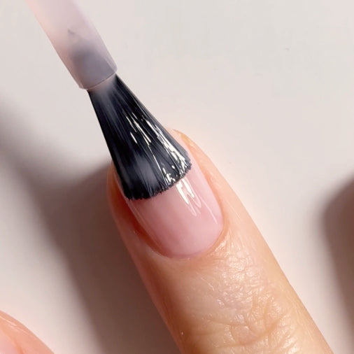A close-up of a hand with a brush applying clear nail polish to a fingernail with a light pink base.