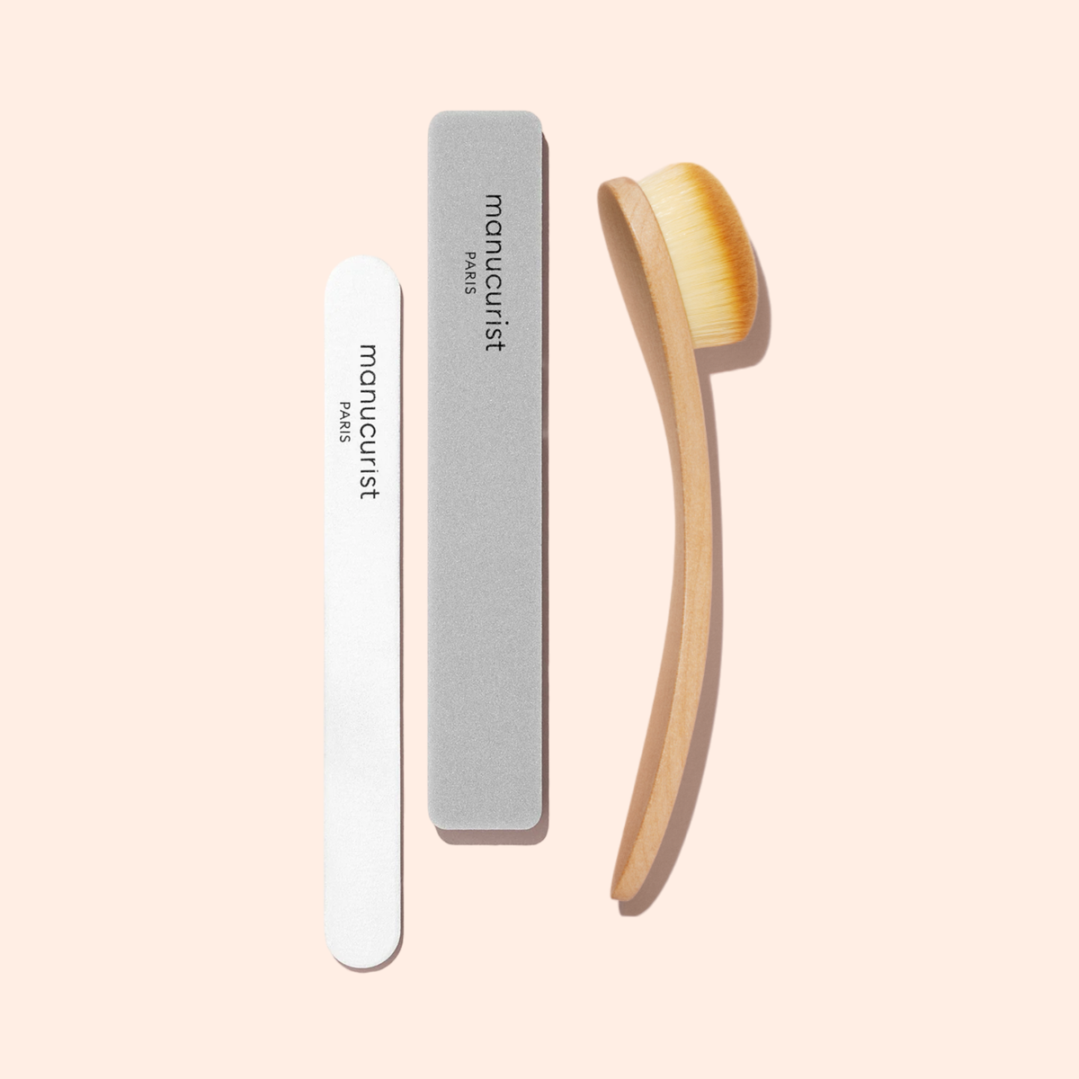 Trois outils de manucure sur un fond pêche clair : une lime à ongles blanche, un polissoir gris et la Dust Brush Manucurist avec des poils véganes et un manche en bois, chacun étiqueté manucurist PARIS.