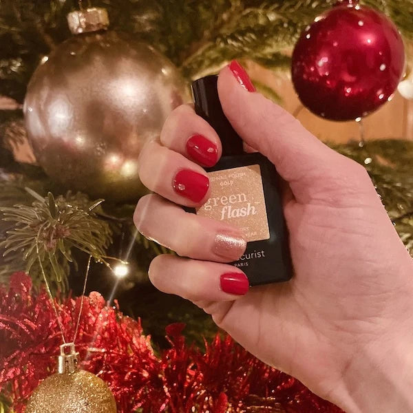 Une main portant un vernis à ongles pailleté rouge et doré tient un flacon de vernis Green Flash devant un sapin de Noël décoré d'ornements dorés et rouges.