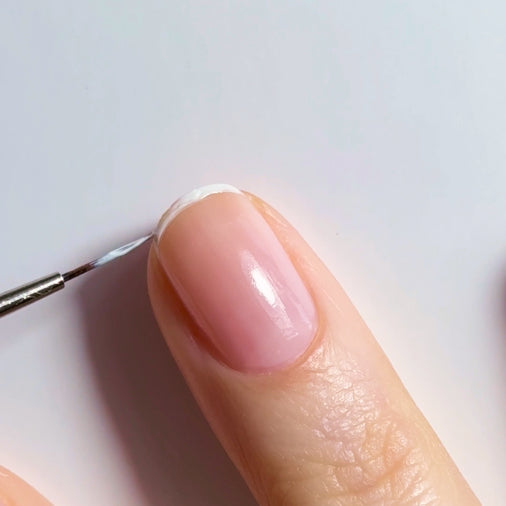 A close-up of a hand with a natural pink nail. A thin brush is applying white nail polish to the tip to create a French manicure.