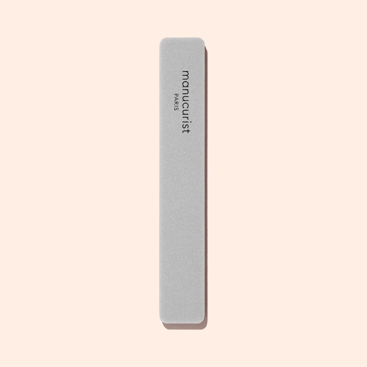 Une éponge polissoir à ongles Manucurist grise rectangulaire aux bords arrondis est placée à la verticale sur un fond beige clair, idéale pour lisser la surface de l&