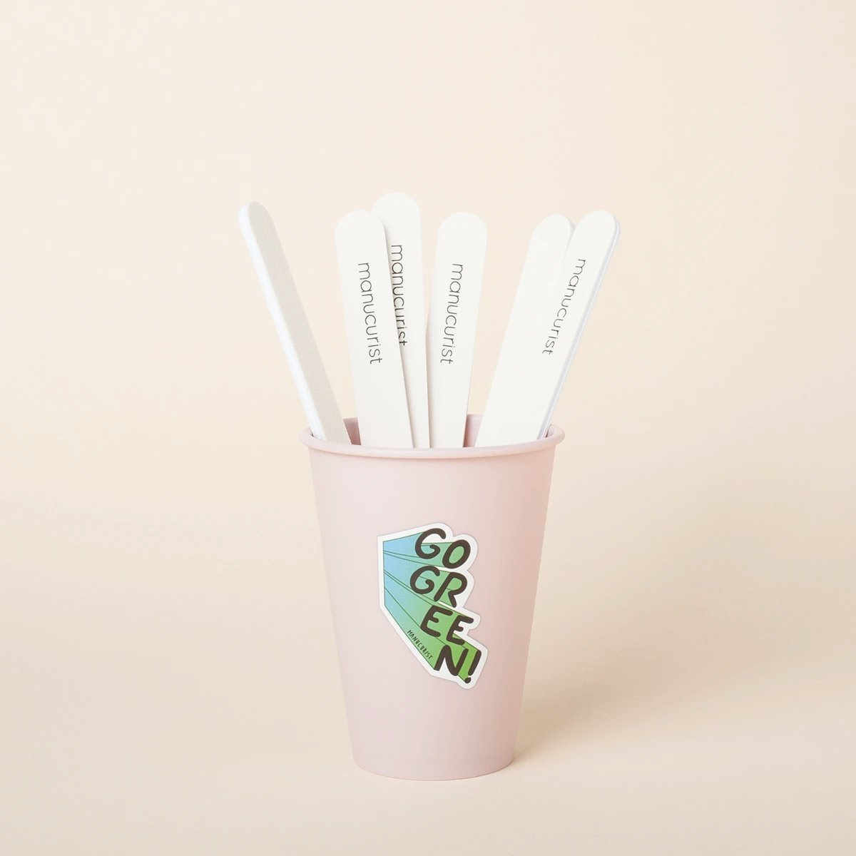 A light pink cup with a “GO GREEN!” sticker holds five white nail files standing upright against a plain beige background. The nail files are labeled manucurist.