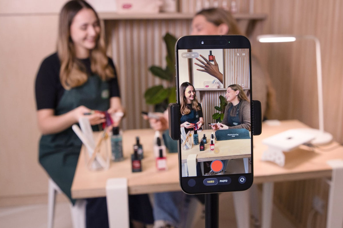 Deux femmes sont assises à une table avec des produits de soin et se filment sur un smartphone, qui les montre en train de parler et de présenter des produits dans une pièce lumineuse et moderne.