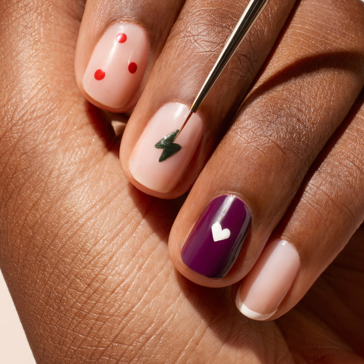 Une main manucurée présente un Nail Art — pois rouges, éclair vert, ongle violet avec un cœur blanc, et ongles naturels avec des pointes blanches et des cœurs — tandis que le Fine Tip Brush Manucurist aux poils synthétiques vegan touche un ongle.