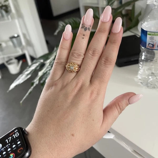 Une main aux ongles longs manucurés rose pâle et portant une bague dorée en forme de fleur est représentée. La main présente de petits tatouages sur les doigts. Une Apple Watch est portée au poignet, et une gourde est posée sur une table blanche à l’arrière-plan.