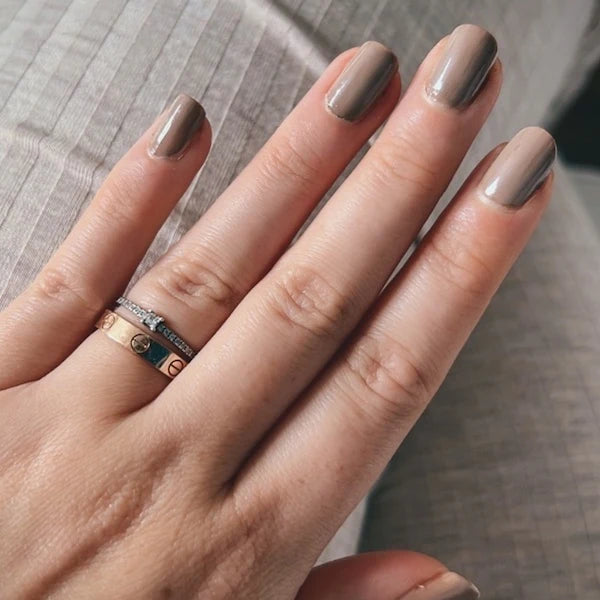 Une main portant un vernis taupe repose sur un tissu gris clair. La personne porte trois bagues superposées — argent, or rose et or — à l'annulaire.