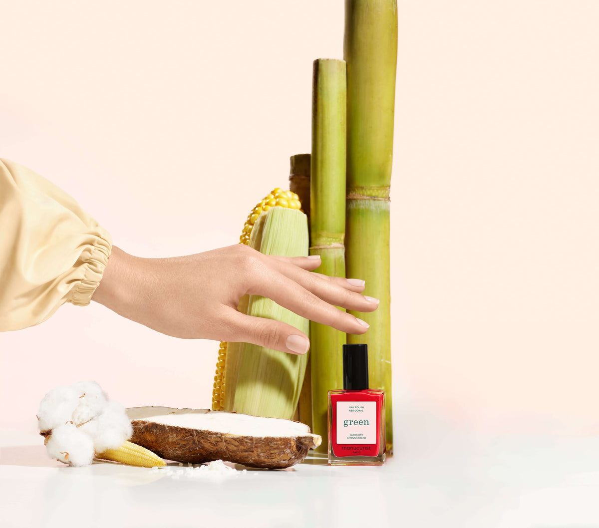 Une main se tend vers un flacon de vernis à ongles rouge portant l’inscription green, à côté de tiges de canne à sucre, d’une tranche de pain avec une tartinade blanche, d’une fleur de coton, de maïs et d’un collier de perles jaunes sur un fond pâle.