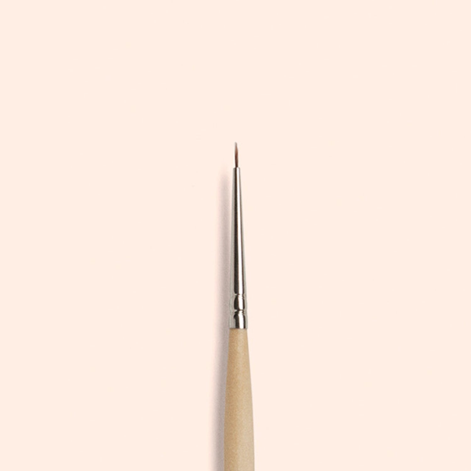 The Manucurist Fine Tip Brush, made with vegan synthetic bristles, a wooden handle, and a silver ferrule, stands vertically against a light beige background—perfect for creating precise nail art.