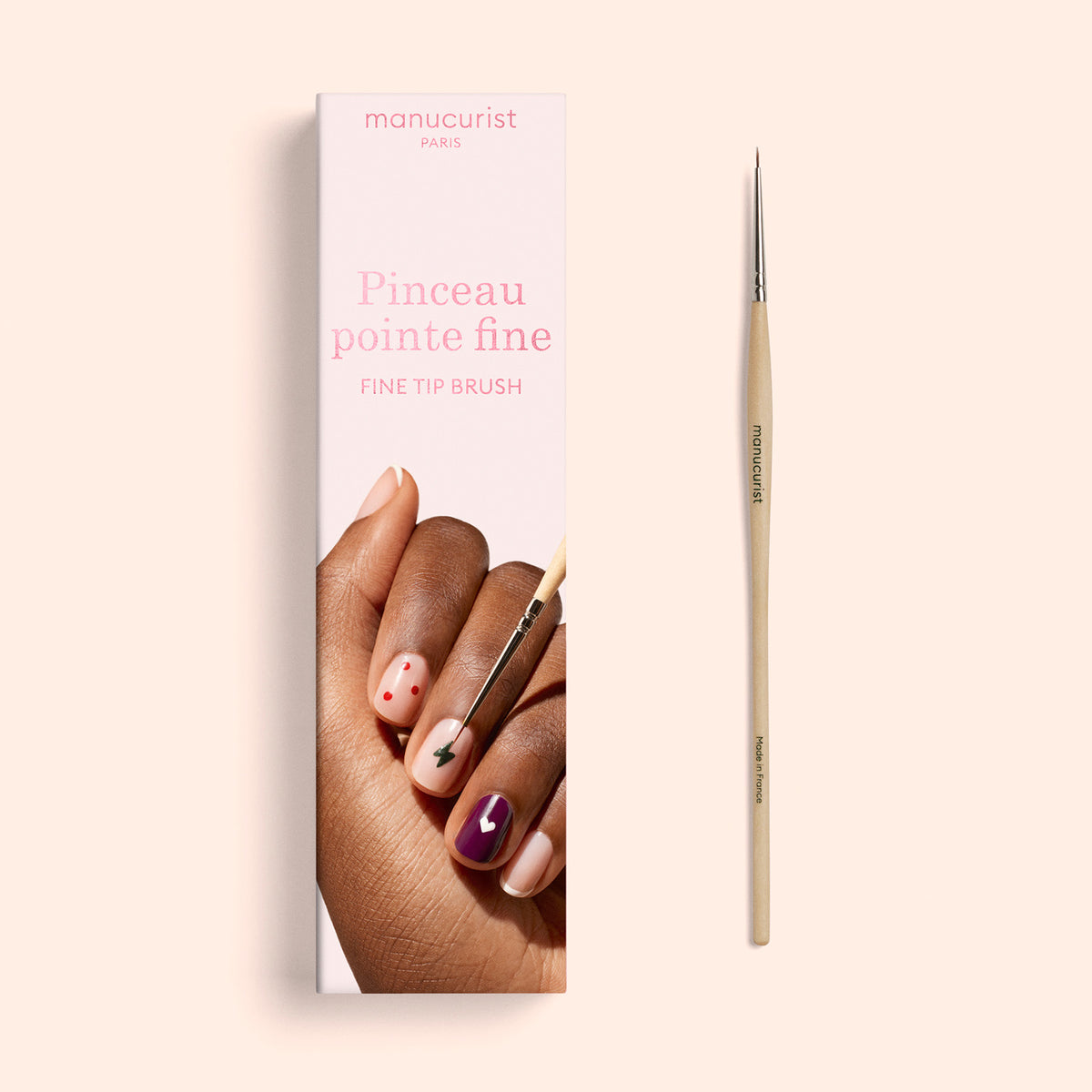 Un Fine Tip Brush de Manucurist aux poils synthétiques vegan est placé à côté de sa boîte, qui présente des images d’ongles peints dans plusieurs couleurs et motifs. L’emballage affiche le texte « manucurist PARIS » et « Fine Tip Brush / Pinceau pointe fine ».