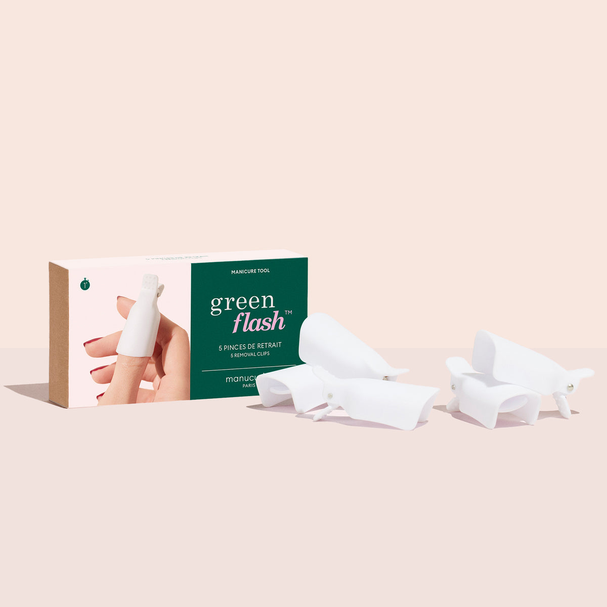 A Manucurist-us Green Flash™ Remover Clips box sits next to five white clips for gentle gel polish removal on a light pink surface, featuring a hand using a clip on the packaging.