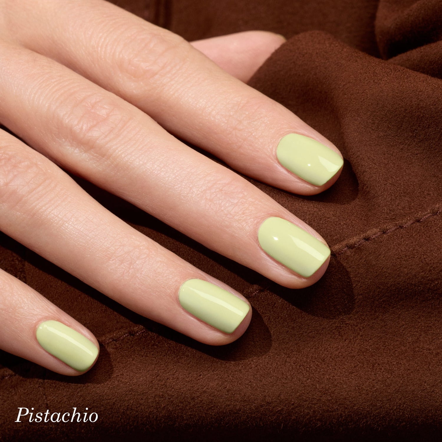 A hand with short, neatly manicured nails painted in a pastel green shade, resting on a brown fabric surface. The word Pistachio appears at the bottom left corner.