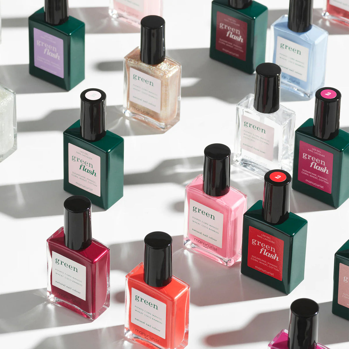 Bottles of nail polish in various colors, including pink, red, green, white, and gold, are arranged on a white surface. Each bottle has a black cap and a rectangular label with the “green flash” brand name visible.