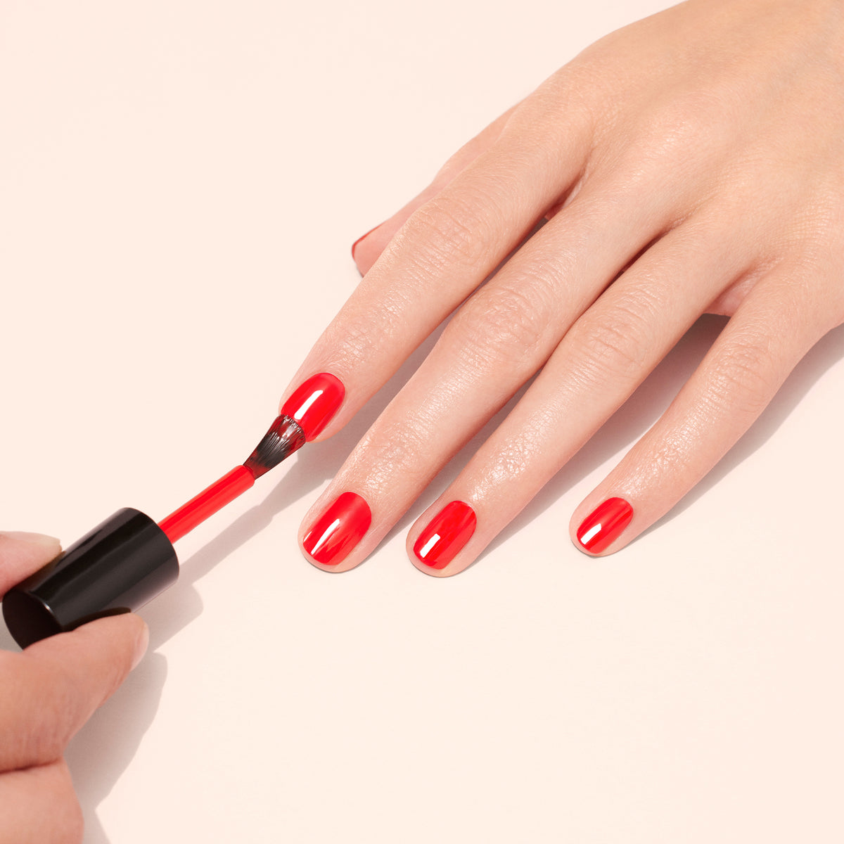 A hand is being painted with bright red nail polish. One hand applies the polish while the other hand rests on a light surface, with glossy red nails partially done.