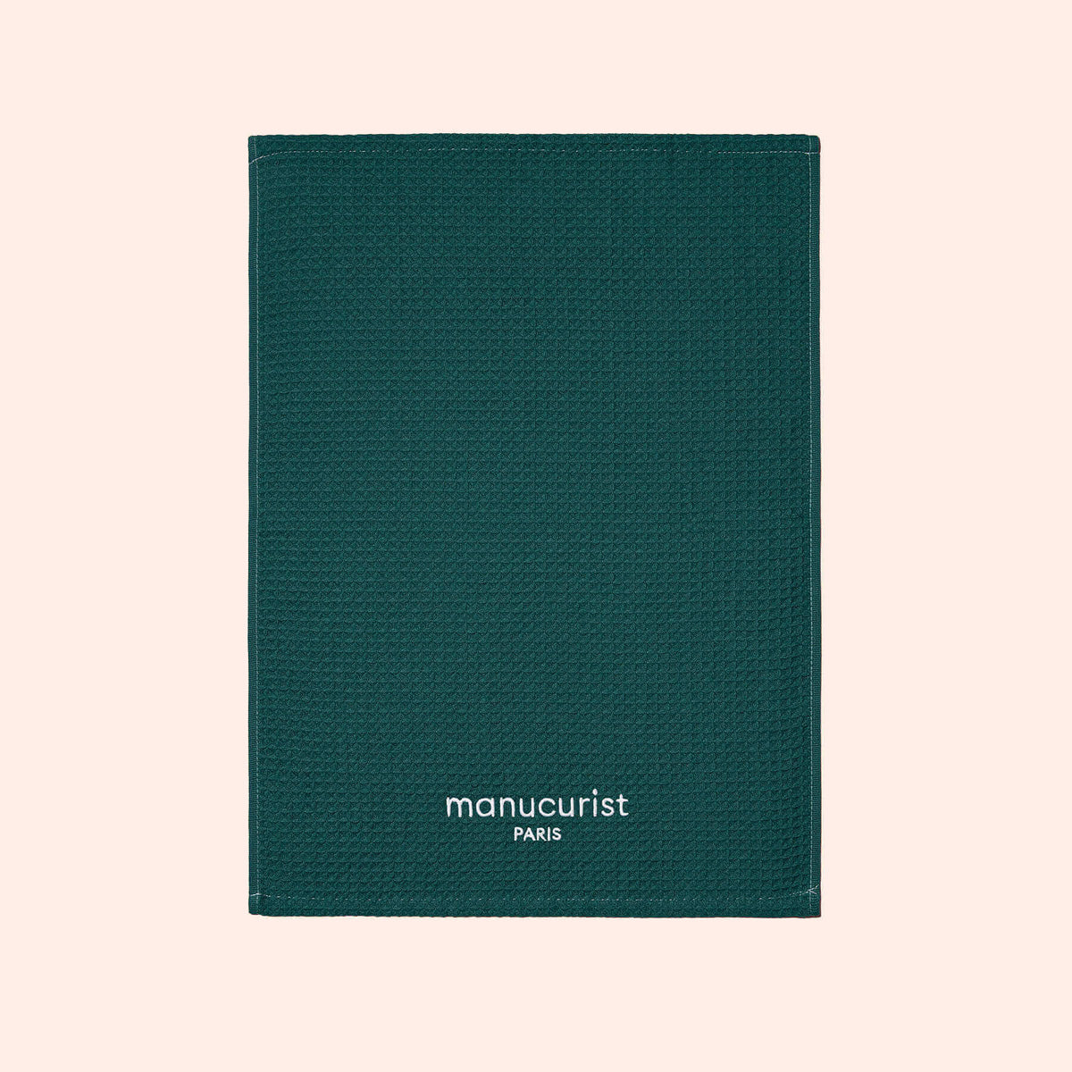 Une serviette verte texturée de Manucurist-us, avec le texte blanc "manucurist PARIS" en bas au centre, présentée sur un fond beige clair et idéale pour protéger les surfaces.