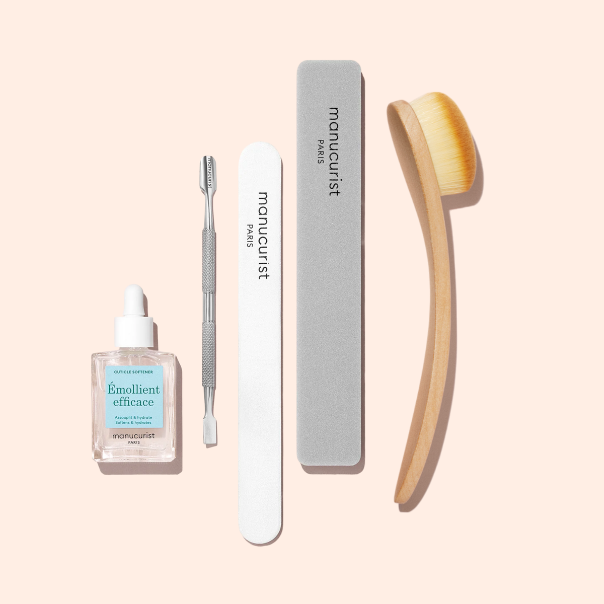 Un ensemble d’outils de manucure sur fond clair, comprenant un émollient pour cuticules, un repousse-cuticules en métal, une lime à ongles blanche, un polissoir gris et la Dust Brush Manucurist à poils véganes.