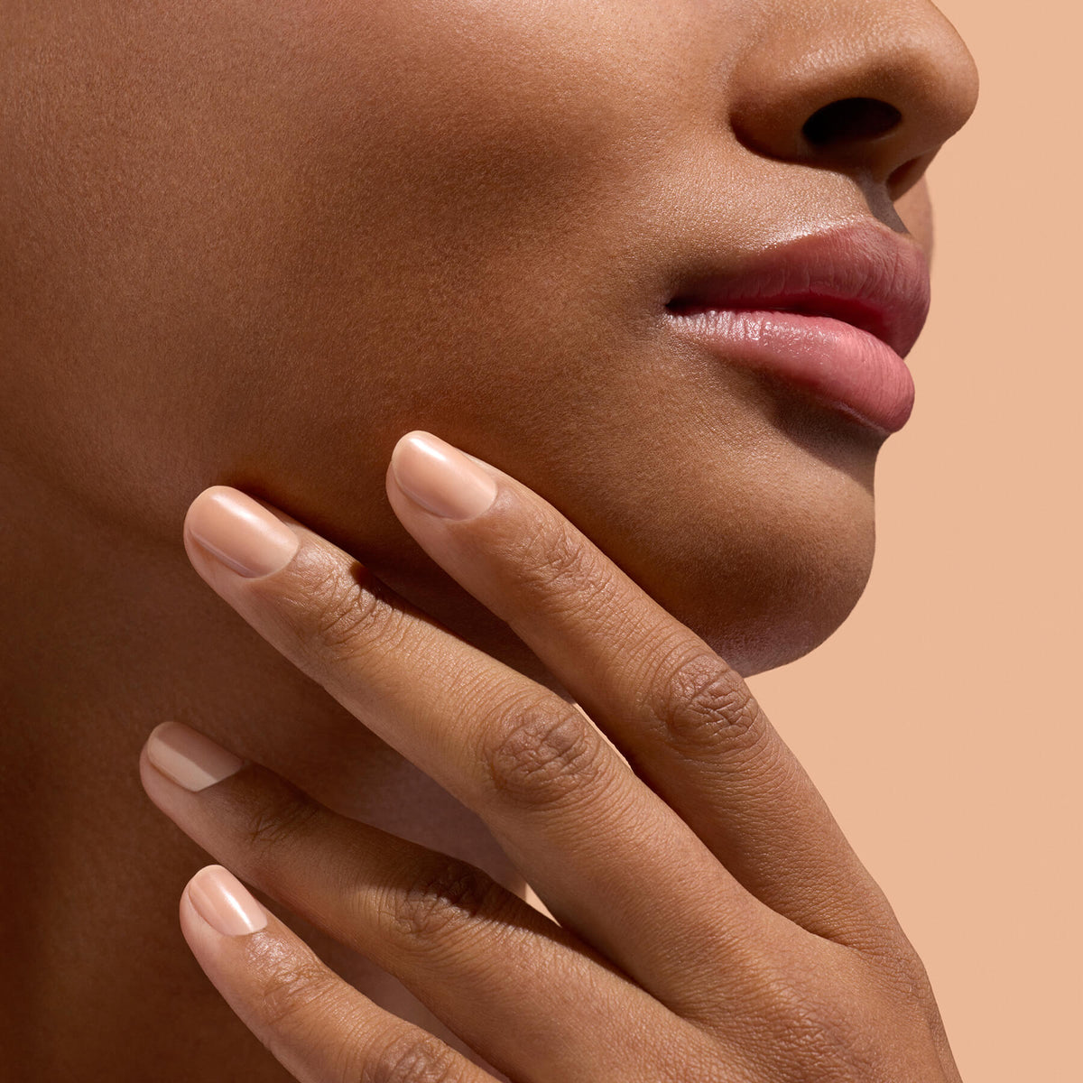 Gros plan sur le bas du visage et la main d’une personne, révélant une peau lisse, des lèvres naturelles et des ongles soigneusement manucurés — mettant en lumière un soin des ongles méticuleux avec Manucurist Active™ Smooth 02 — sur fond beige.