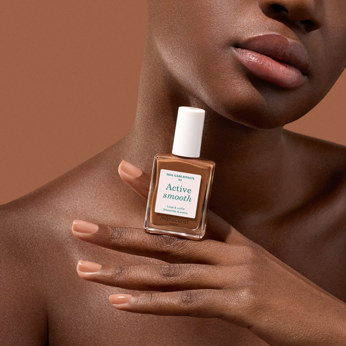 Une personne à la peau brune et lisse tient un vernis à ongles Manucurist Active Smooth 03 près de son visage, dévoilant des ongles manucurés nude sur un fond brun assorti pour mettre en valeur les bienfaits d’un soin des ongles de qualité.