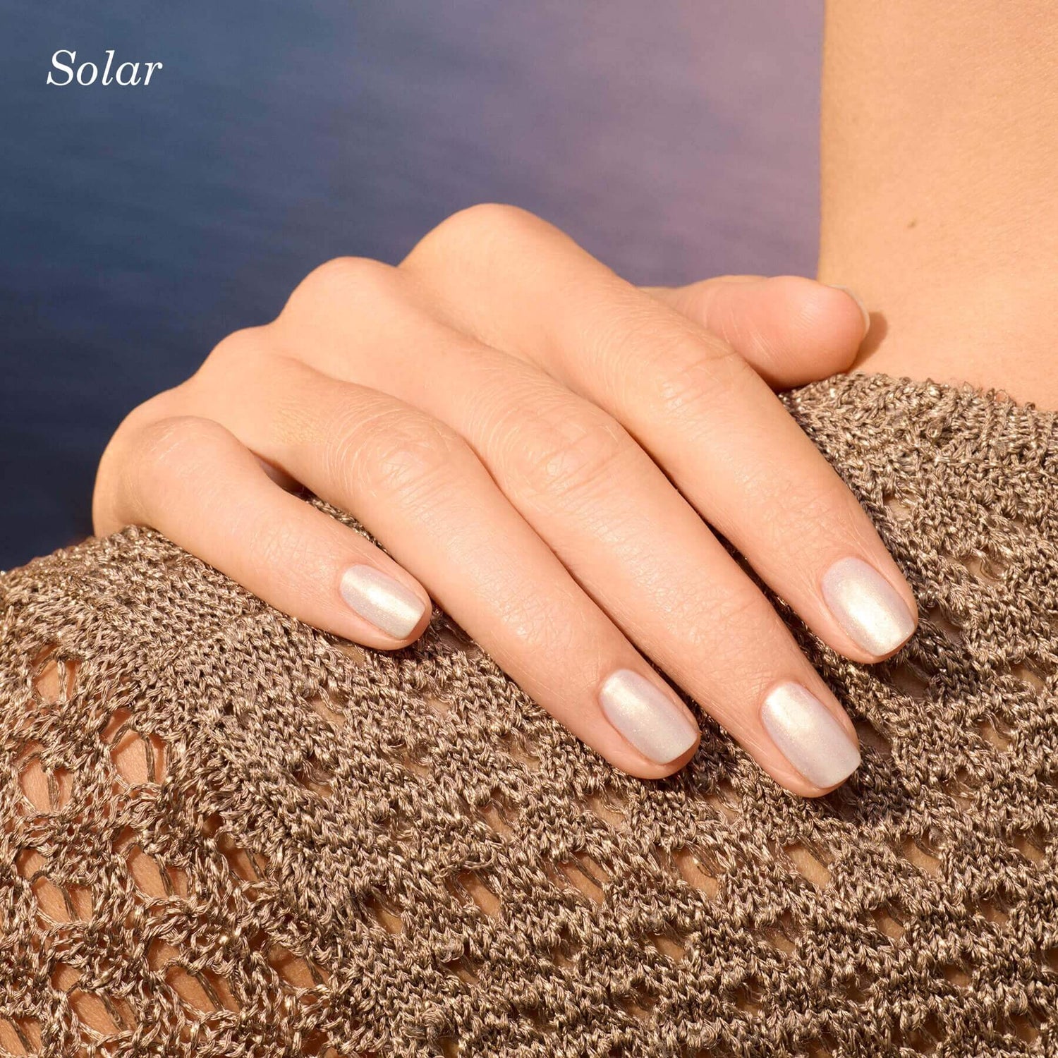 A close-up of a hand with short, pearly white nails resting on a shoulder covered by a textured, brown knit top. The background is softly blurred in blue and purple hues. The word Solar is in the top left corner.