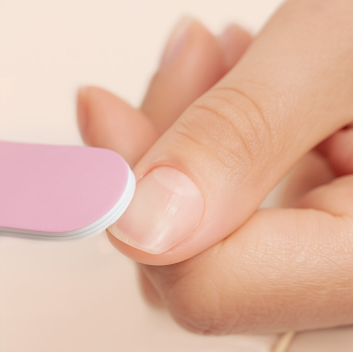 Close-up of a person using a pink nail file to shape their fingernail, with a focus on the hand and nail. The background is soft and neutral.