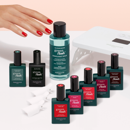 A hand with red polished nails reaches toward the Manucurist-test Green Flash™ Welcome Kit, featuring gel polish, acetone-free remover, clips, and a UV nail lamp on a light pink surface.