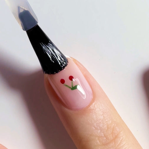 A close-up of a finger with light pink nail polish featuring a small cherry design. A brush is applying a clear top coat over the nail art.