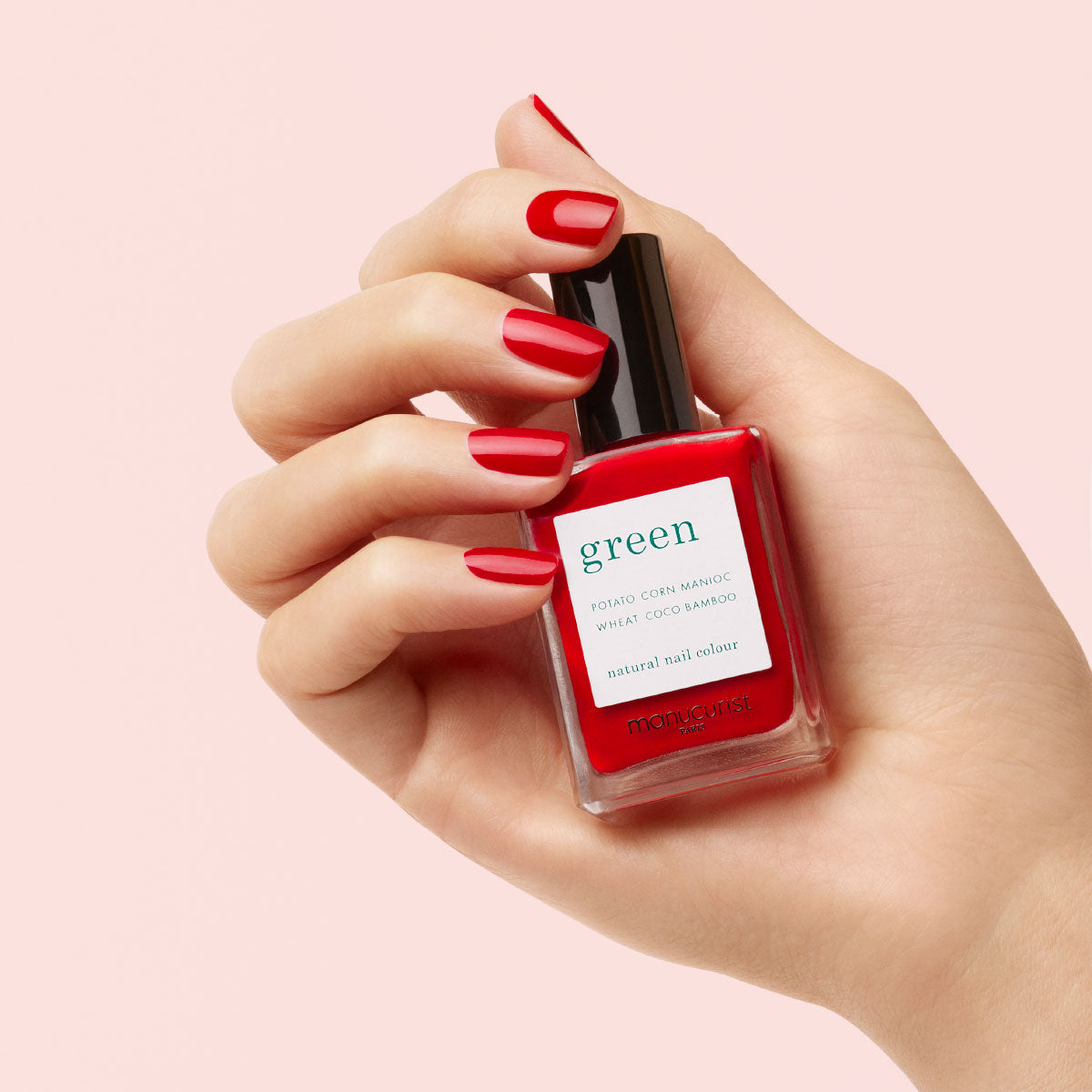 A hand with neatly manicured red nails holds a bottle of red nail polish labeled “green” by Manucurist against a light pink background.