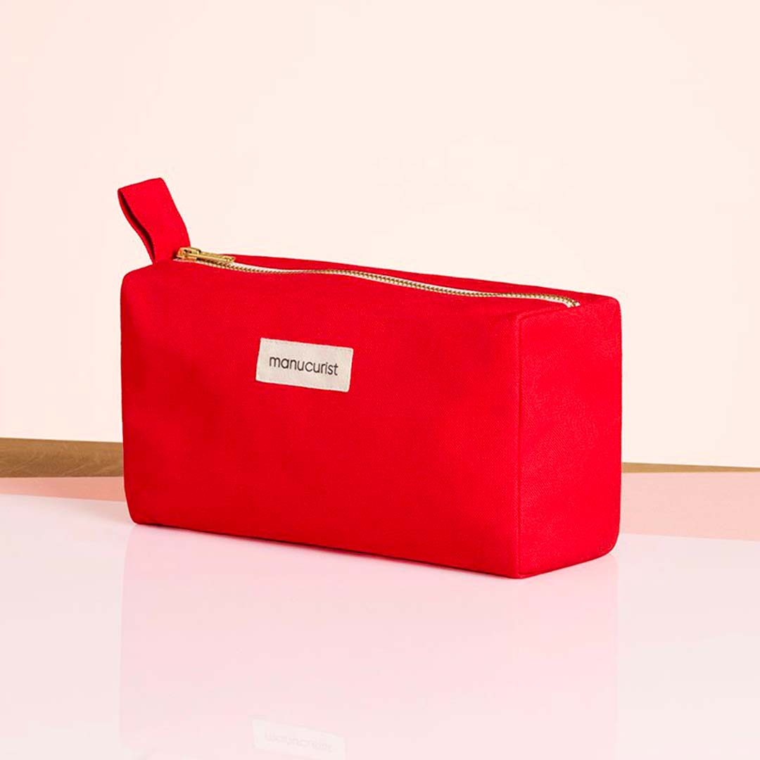 Le Red Travel Bag Manucurist-us, une pochette rectangulaire en coton biologique avec fermeture éclair et poignée latérale, est posé sur une surface brillante. Une étiquette blanche avec le nom de la marque est visible. L&