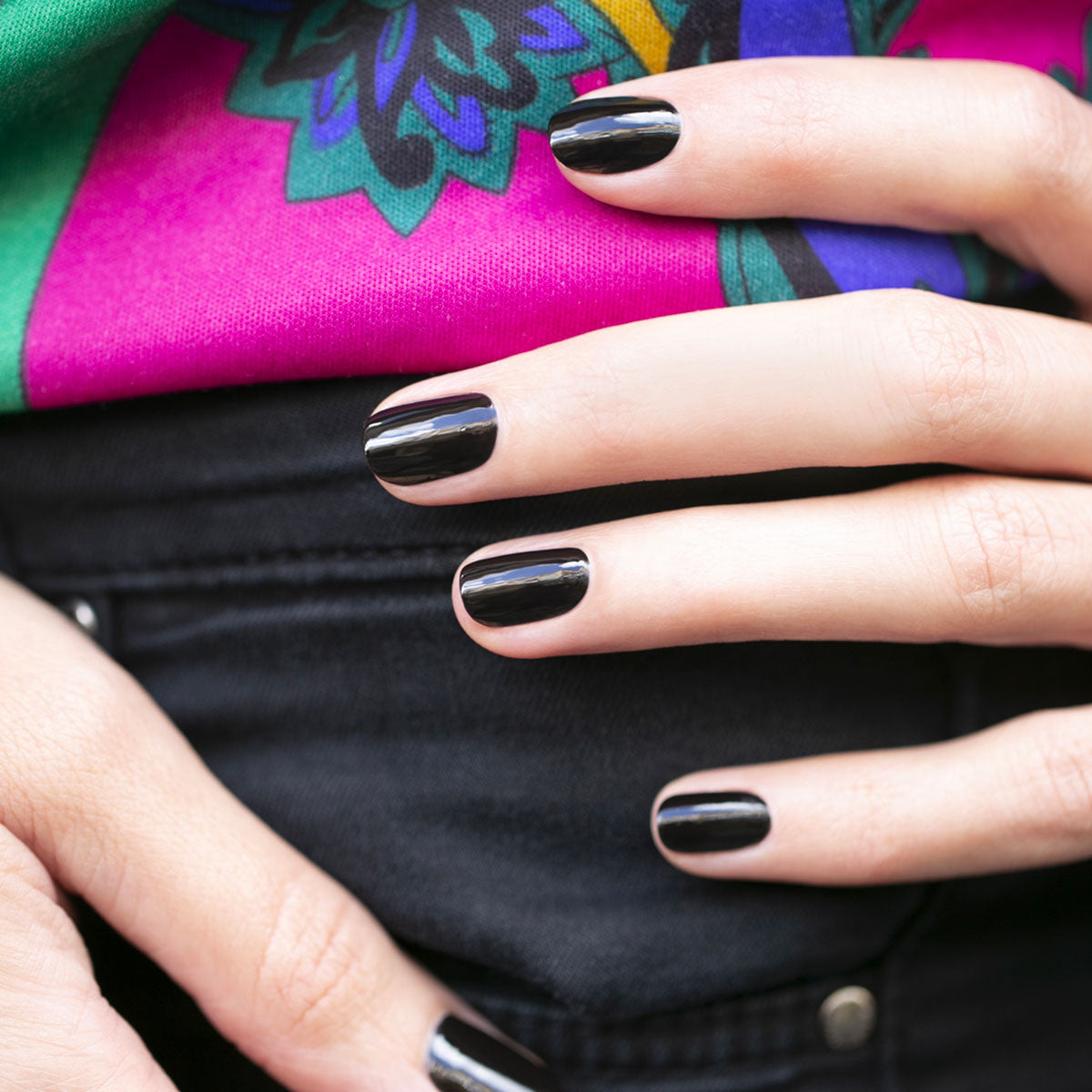 Gros plan d’une main avec le vernis Licorice de Manucurist de la gamme Green™, posée sur un jean noir devant un fond à motifs rose vif et vert.