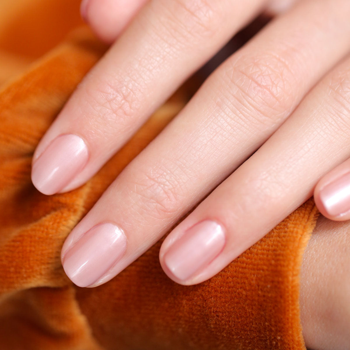 Gros plan d’une main aux ongles soigneusement manucurés, peints en Carnation par Manucurist, posée sur un tissu en velours orange.