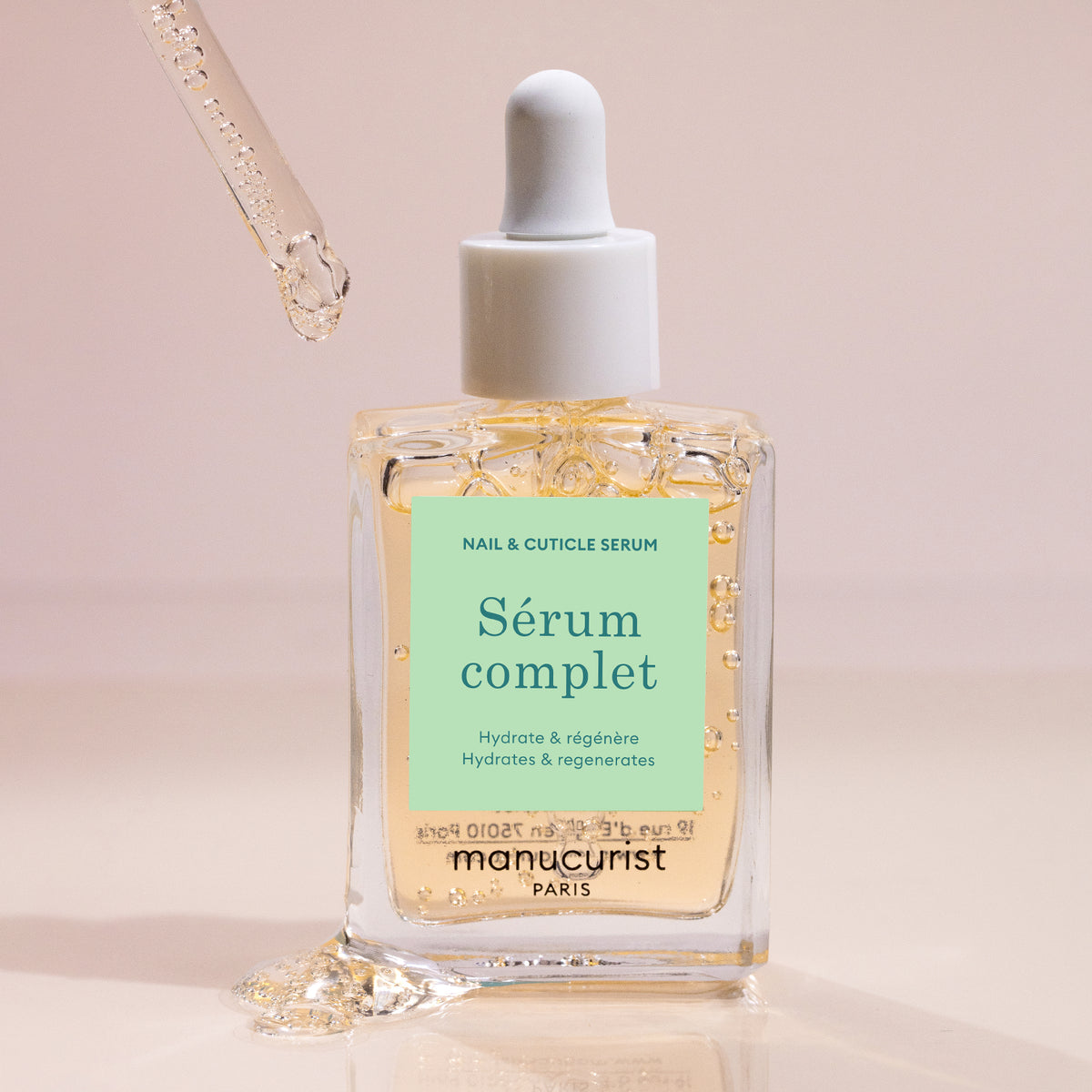 Une pipette dépose du Sérum Hydratant Manucurist dans un flacon en verre transparent portant le nom de la marque. Le fond beige doux met en valeur les gouttes de ce soin nourrissant pour les ongles visibles sur le flacon.