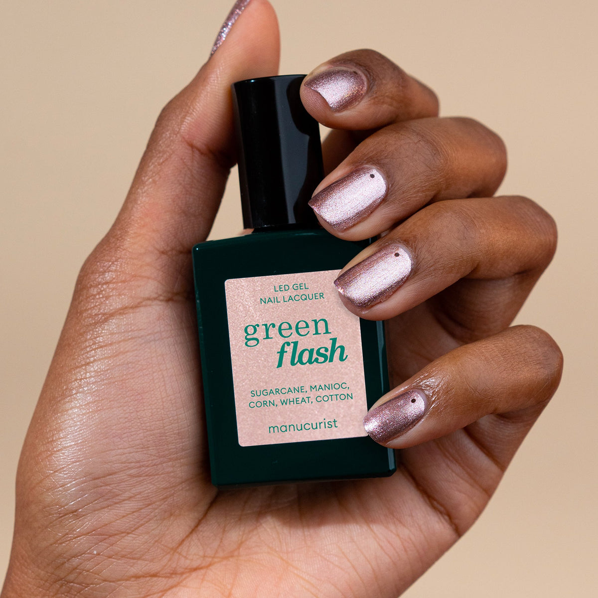 A hand with iridescent pink nail polish holds a bottle of Manucurist Cosmic Rose gel nail lacquer against a beige background.