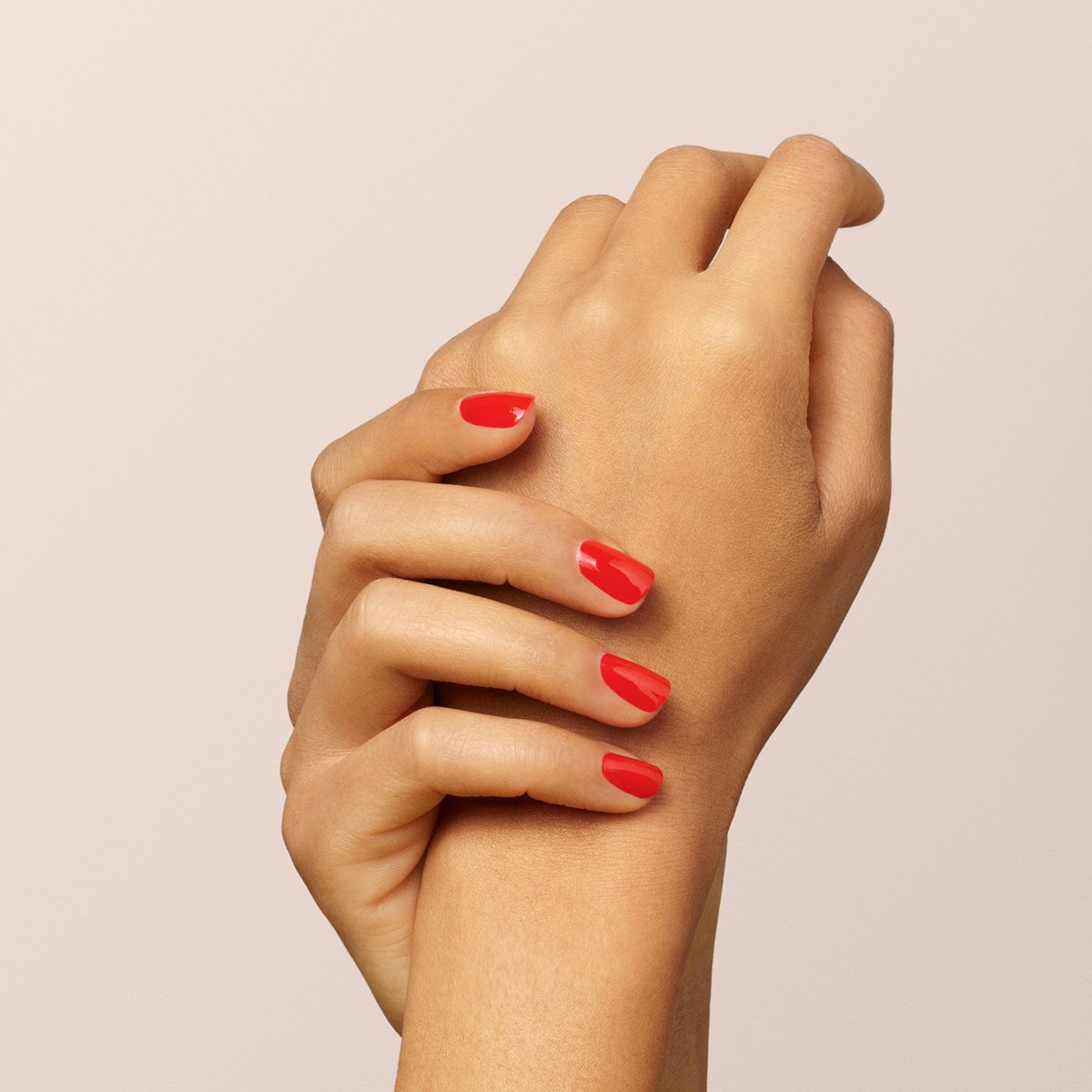 Two hands with neatly manicured nails painted in Manucurist’s Red Coral nail polish are gently clasped together against a soft beige background.